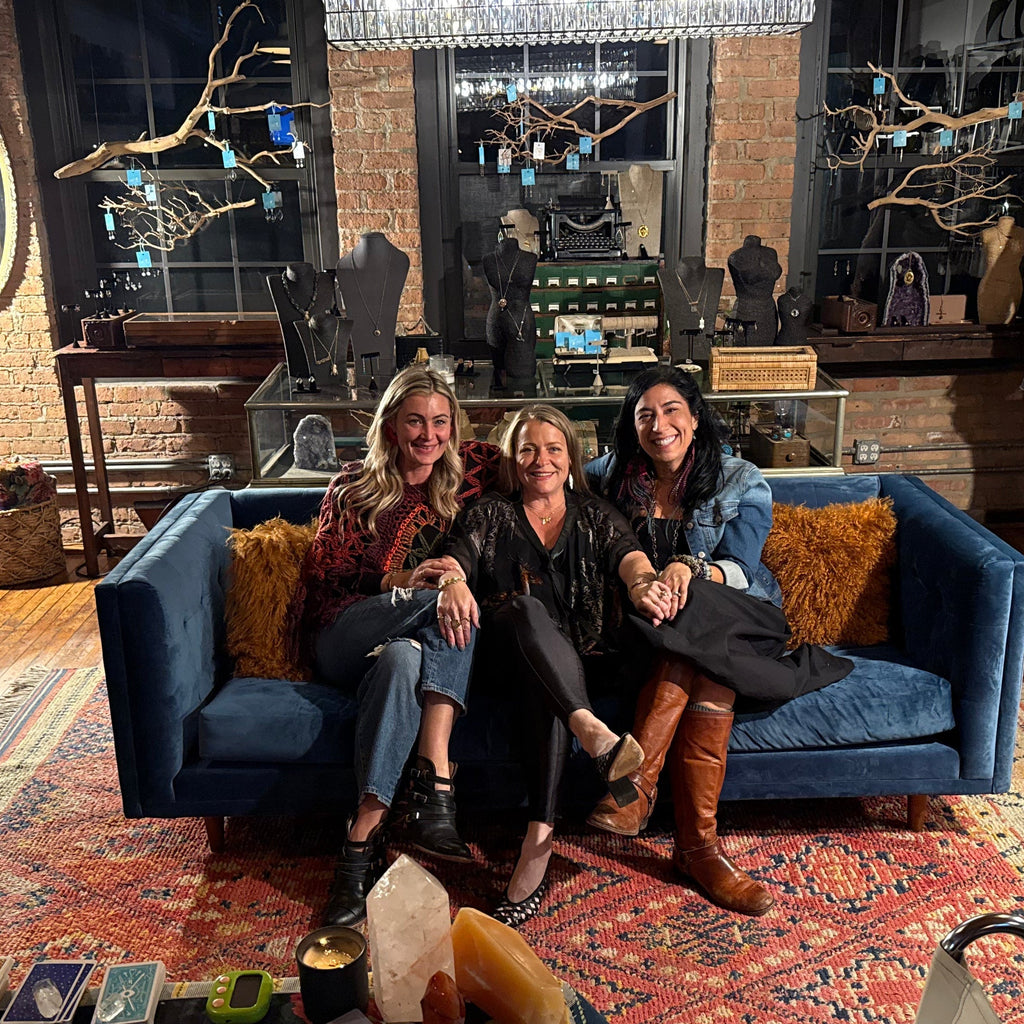 Three women sitting together on a blue velvet sofa in a cozy, rustic boutique showroom with brick walls, wooden floors, and shelves of handbags and accessories.
