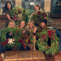 Five women posing together in a cozy indoor setting, each holding a large, festive green holiday wreath decorated with red and white accents. They are seated and standing behind a blue couch with a vintage trunk in the foreground.