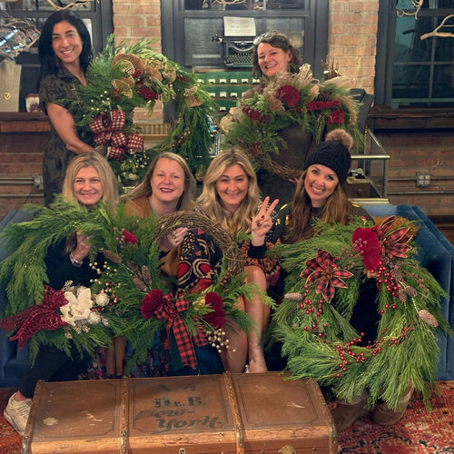Five women posing together in a cozy indoor setting, each holding a large, festive green holiday wreath decorated with red and white accents. They are seated and standing behind a blue couch with a vintage trunk in the foreground.